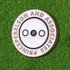A round, soft enamel magnetic ball marker in gold and white with a central black dot, set against a grassy background.