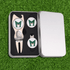 A silver gift box containing a metal golf divot tool and two golf ball markers with a logo. The background is green grass.