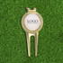 A gold golf divot tool with a circular ball marker, resting on green grass. The marker features a logo.