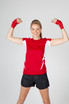 A woman in a red and white short-sleeved T-shirt flexes her arms, wearing black shorts and red hand wraps. A woman in a red and white short-sleeved T-shirt flexes her arms, wearing black shorts and red hand wraps.