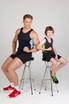 A man and boy sit on stools, both wearing black singlets with contrasting blue and green accents, showing off their muscles.
