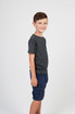 A grey heather t-shirt is worn by a young boy standing sideways, paired with navy shorts. The shirt has a logo.