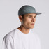 A five-panel cap in a soft blue-grey colour worn by a model, featuring a subtle logo.