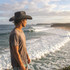 A person wearing a black straw hat stands by the ocean, gazing at the waves and surfers.