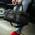 A black duffle bag with a logo, held by a person in casual jeans and red sneakers.