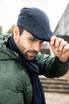 A man wearing a dark blue cap and a green jacket, adjusting his cap with a serious expression.