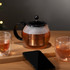 A glass tea press with a black handle, filled with brewed tea, alongside two cups and a smartphone on a wooden surface.