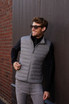 A man wearing a grey down vest stands against a brick wall, smiling and wearing sunglasses. The vest has a logo.