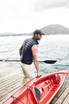 A man wearing a navy down vest and a cap, carrying a red kayak by a waterfront.