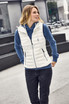 A white down vest worn over a blue hoodie, displayed against an outdoor modern café setting. The vest features a front zip and pockets.