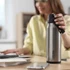 A drink bottle in stainless steel with a black cap, featuring a logo, placed on a desk next to a notebook and phone.