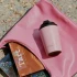 A pink reusable cup resting on a pink pouch, with colourful items visible underneath. The cup has a logo.