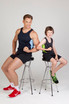 A navy and green singlet worn by an adult and a child, both seated on stools, showcasing their muscles.