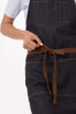 A denim apron in dark blue, featuring pockets and a brown tie belt, with a hand adjusting the waist strap.