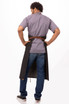 A dark grey bib apron worn by a person facing away, with a brown belt, styled over a grey shirt and blue jeans.