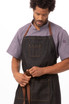 A dark denim bib apron with leather straps, worn by a person adjusting the waist tie. Featuring a logo.