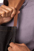 A close-up of a person holding the brown strap of a dark denim chef's bib apron. The background is neutral.