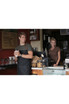 A dark grey Memphis Chefs Bib Apron worn by a man holding a drink in a café setting, with a woman in a brown shirt nearby.