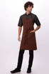 A brown Dorset Bib Apron worn by a model, featuring a pocket and tied at the waist. The model also wears a dark shirt and pants.