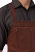 A brown denim bib apron with a front pocket, worn over a black shirt. The apron features metal fasteners.