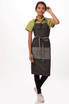 A woman in a green shirt and black Manhattan bib apron stands against a white background. The apron features a front pocket and ties.