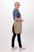 A woman wearing a beige Logan Bib Apron over a blue shirt, featuring pockets and a tied back.