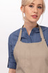 A beige bib apron with a pocket is worn by a model, featuring a blue striped shirt underneath. The image has a logo.