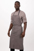 A man wearing a grey bib apron over a collared shirt, standing against a plain background. The apron has a logo.