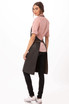 A stylish black bib apron worn over a pink shirt, featured on a model with long hair. Includes a logo.
