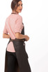 A woman wears a black Boulder Bib Apron over a pink shirt, facing slightly away from the camera.