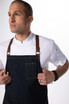 A denim cross back apron in dark blue with brown straps, worn over a white shirt by a model.
