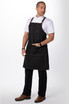 A man stands wearing a black denim cross back apron over a white shirt and black trousers, featuring pockets and a branding logo.