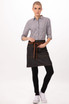 A woman wearing a stylish black and denim half bistro apron with a brown tie, paired with a checkered shirt and black pants.
