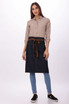 A denim bistro apron in navy with a brown waist tie, worn over a beige shirt and blue jeans. Features branding.