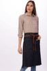 A woman models a dark denim bistro apron with brown accents, featuring pockets and a tie at the waist.