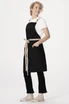 A black cross back apron with a beige belt and pockets, worn by a model with curly hair.