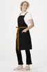 A black Lockharte cross back apron featuring a brown tie waist and pockets, worn over a white t-shirt.