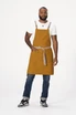 A man wearing a mustard-coloured cross back apron with a grey tie, denim jeans, and sneakers. The apron has a logo.