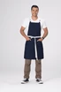 A man wearing a navy blue Lockharte Cross Back Apron with a white belt, standing confidently with hands in pockets.