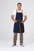 A cross back apron in navy with tan straps and a waist tie, featuring a model standing with hands in pockets.