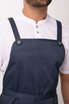 A navy blue bib apron features adjustable straps and a front pocket, worn over a white shirt. It has a logo on it.