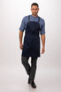 A navy blue bib apron with adjustable straps, displayed on a model wearing a denim shirt and smart pants.