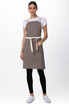 A woman wears a brown Byron Cross-Back Apron with a cream strap, paired with a white top and black pants.