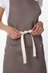 A brown cross-back apron with a cream tie, featuring a pocket at the front and a logo.