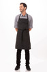 A black bib apron with contrasting ties worn by a smiling man, standing upright with hands behind his back.