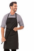 A man in a black bib apron with contrasting ties, smiling while holding a tie. The apron features a logo.