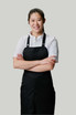 A person wearing a black bib apron over a white shirt, arms crossed, smiling against a neutral background.