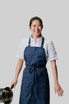 A smiling individual wearing a navy blue Brio Bib Apron, holding a pot, with a logo on the apron.