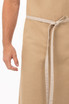 A beige bib apron with a tie waist, featuring a hand reaching towards it.