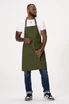 An olive green bib apron is worn by a man, paired with a white t-shirt and blue jeans. The apron has adjustable straps and a logo.